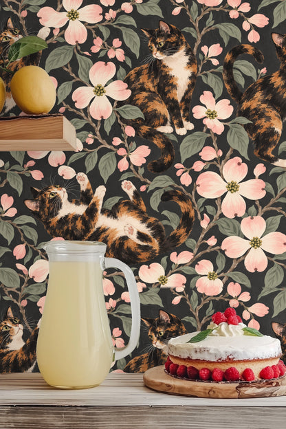 Playful calico cat lying on back with paws raised among dogwood branches on wallpaper.