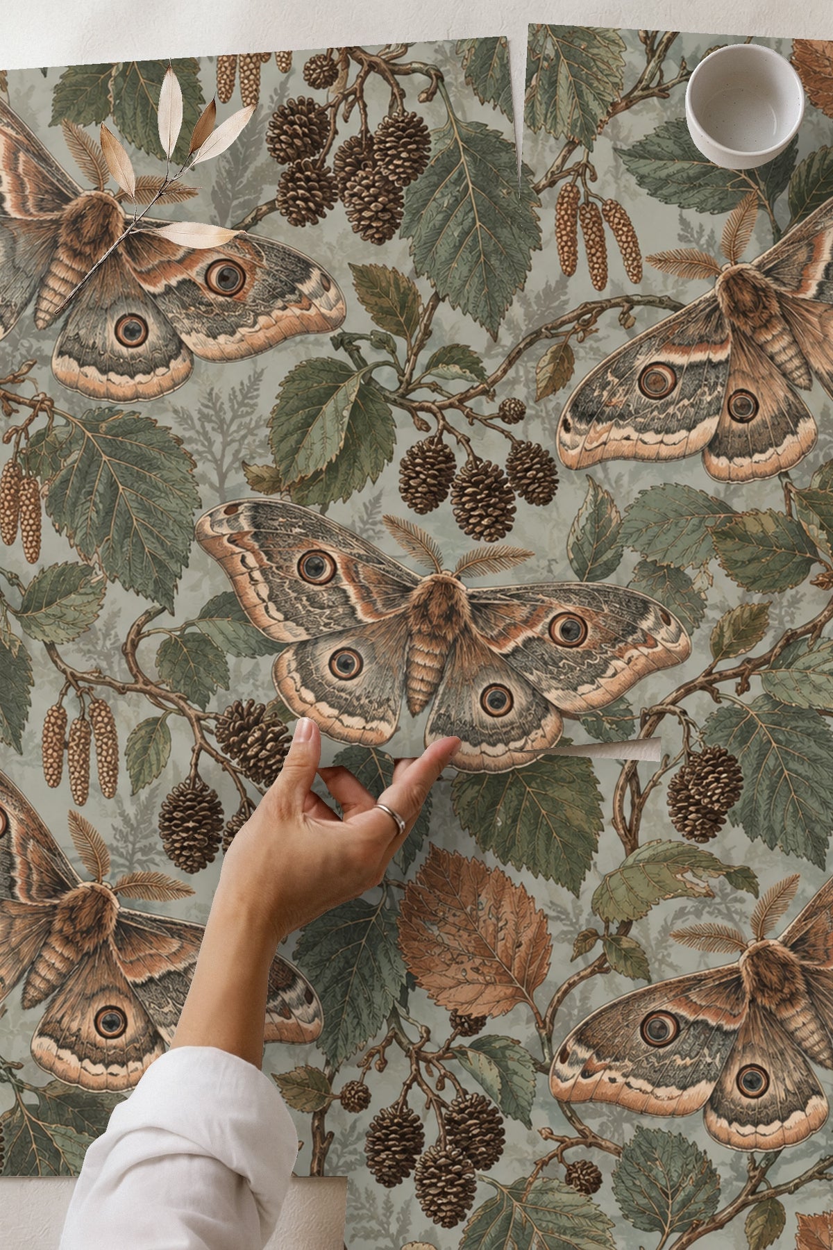 Living room wall covered with Mist Alder Moths wallpaper, featuring a sofa with rust and pink pillows and green velvet pouf.