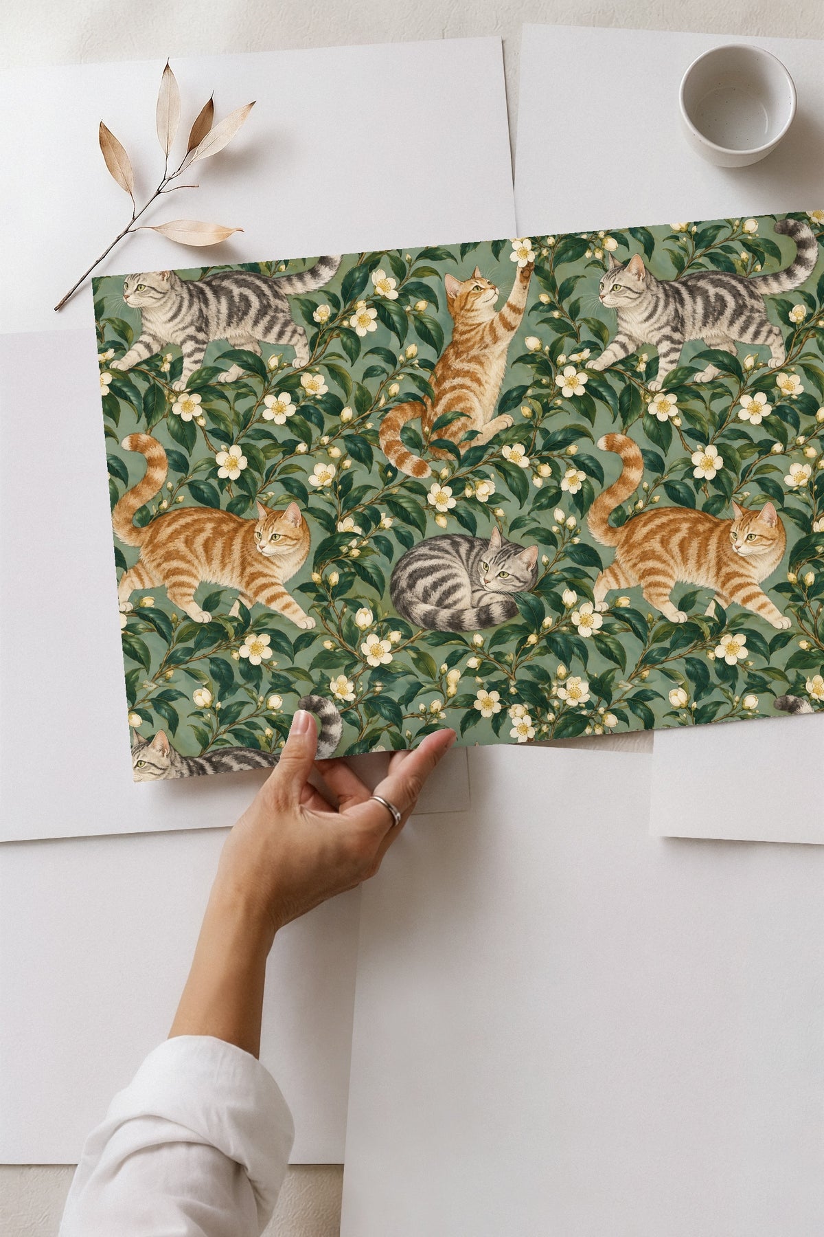 Seated gray tabby cat resting amid camellia leaf clusters and white flowers on wallpaper panel.