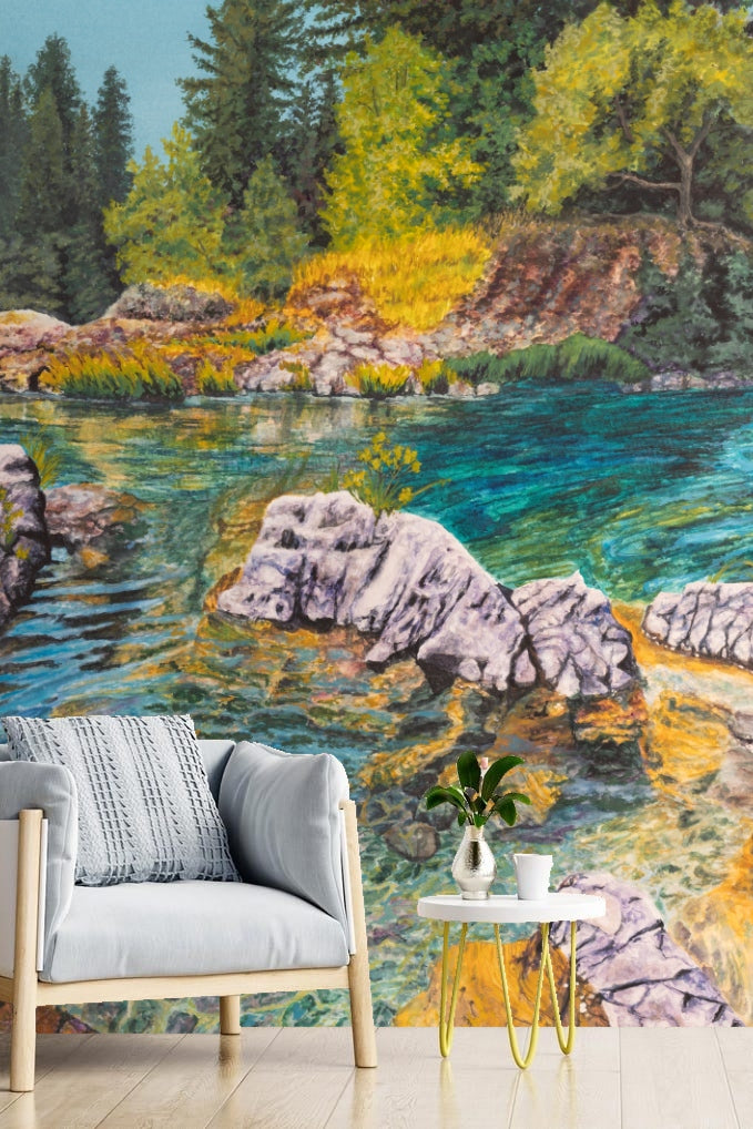 Bedroom interior with sunlit riverbank mural wallpaper behind bed, light wood furniture and neutral bedding.